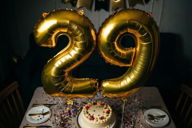 Grote glanzende gouden folieballonnen vormen 29 boven een feesttafel met gekleurde confetti en een verjaardagstaart in een donkere feestelijke omgeving.