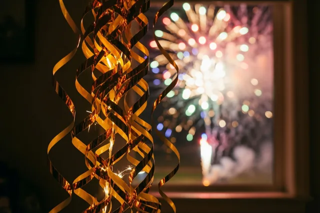 Gouden metallic slingers krullen op de voorgrond en vangen het licht van kleurrijk vuurwerk dat door een raam zichtbaar is.