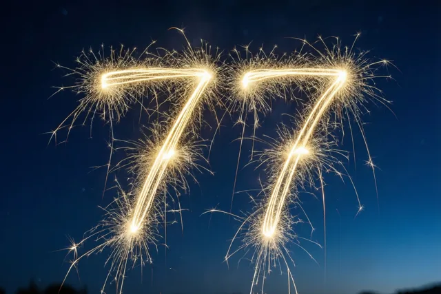 Gouden lichtsporen van sterretjes vormen het getal 77 tegen een donkerblauwe nachthemel.