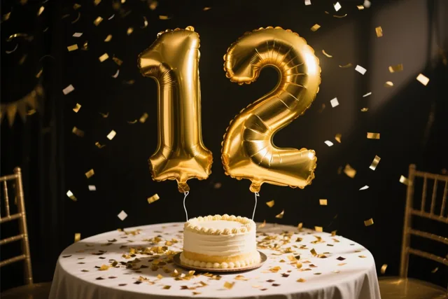 Grote goudkleurige folieballonnen vormen 12 boven een tafel met een wit kleed, gouden confetti en een kleine wit geglazuurde taart, tegen een donkere achtergrond met vallende confetti.