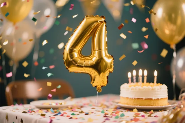 Een grote gouden folieballon in de vorm van een 4 zweeft boven een tafel met kleurrijke confetti, met taart, kaarsen en andere ballonnen tegen een donkere achtergrond.