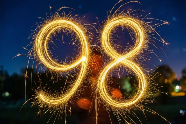 Gouden lichtsporen van sterretjes vormen 98 in de nacht, met onscherpe lichtjes en bomen op de achtergrond.