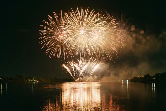 Goud en wit vuurwerk boven een brede rivier weerspiegelt in het water, met bomen en zwakke lichtjes langs de oevers onder een donkere hemel.