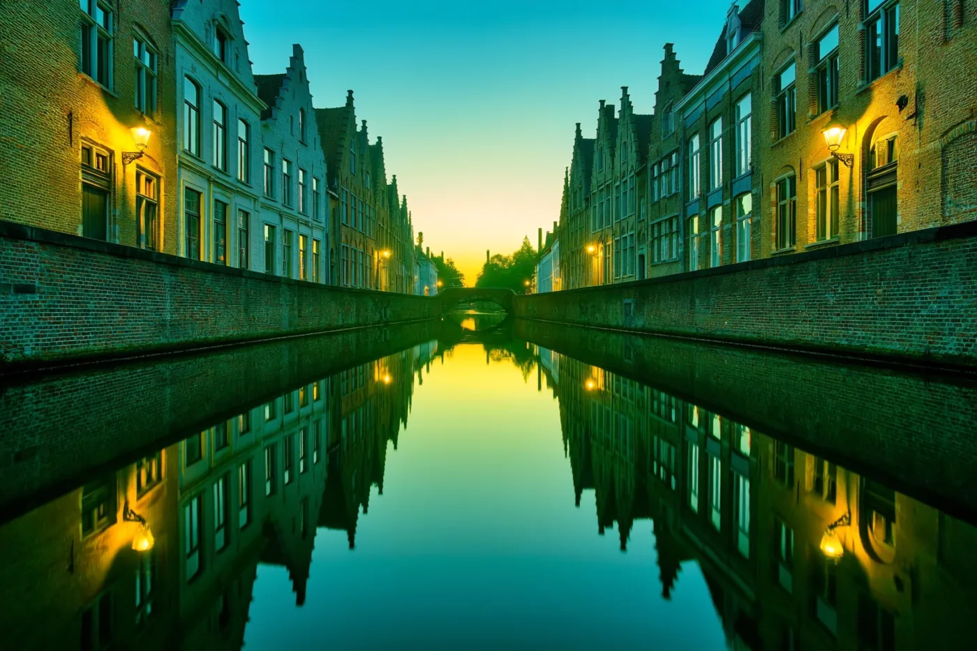Een kanaal in een oud Vlaams stadje ligt bij zonsopgang tussen historische huizen met puntdaken en warme straatlampen, met goudgele weerspiegelingen in blauwgroen water.