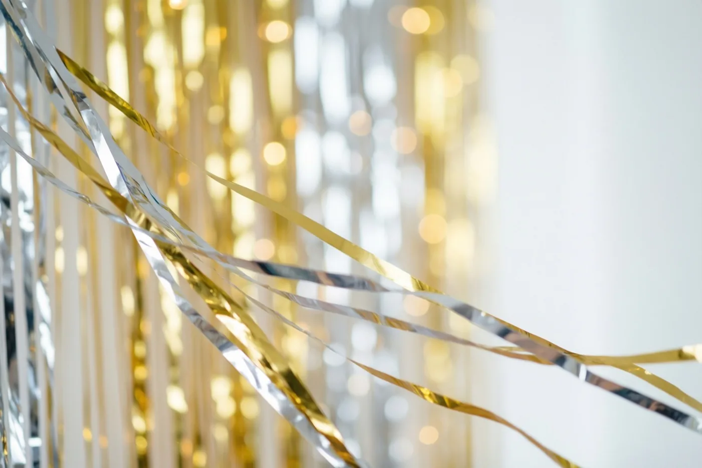 Gouden en zilveren metallic slingers hangen op de voorgrond tegen een achtergrond van wazige lichtjes in goud, zilver en wit.