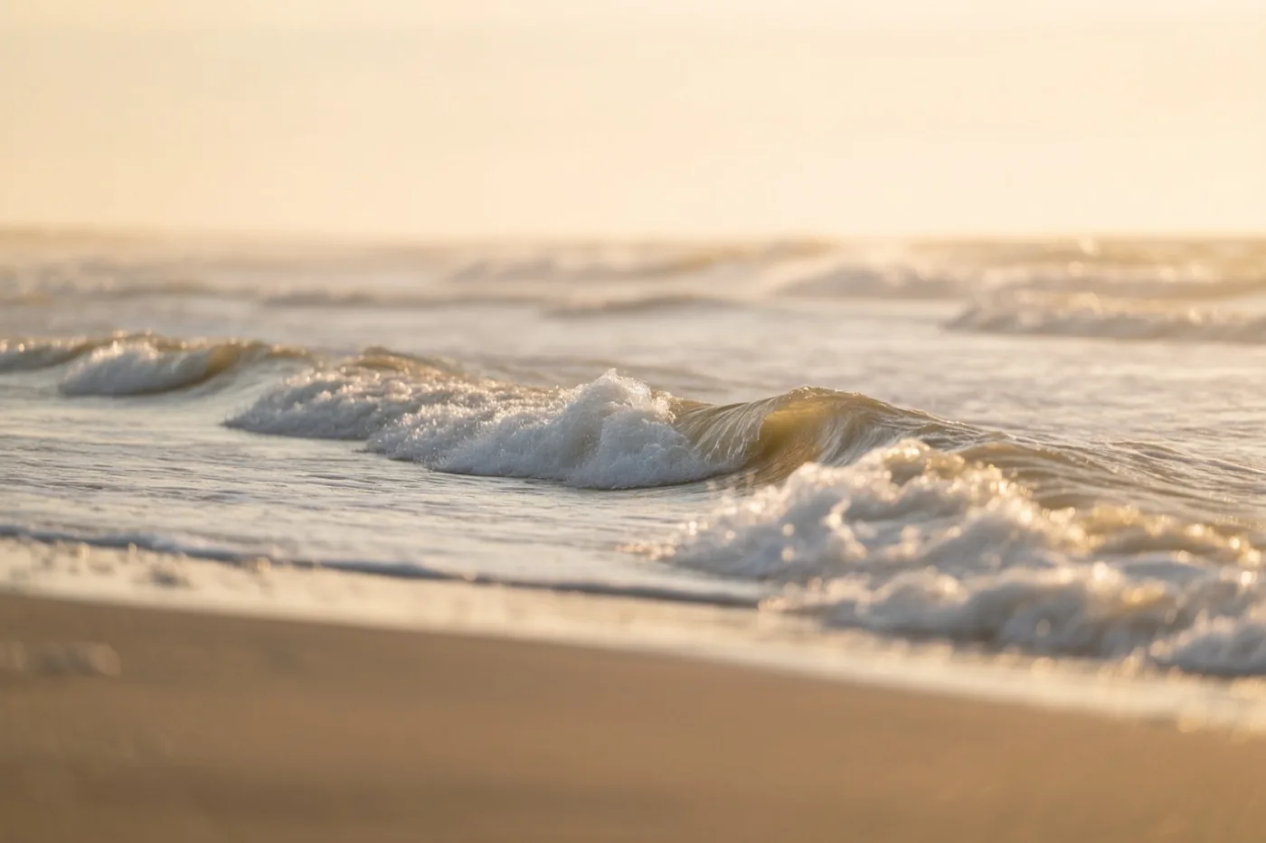 Zachte golven rollen op een zandstrand in warm goudkleurig zonlicht, met beige en witte tinten.