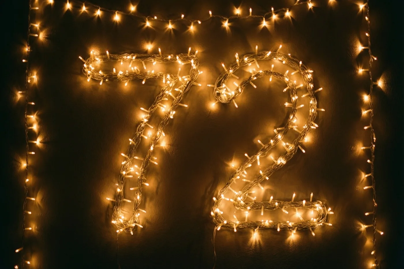 Warme lichtjes vormen 72 op een donkere muur, met goudgele, onscherpe lichtpunten rondom.