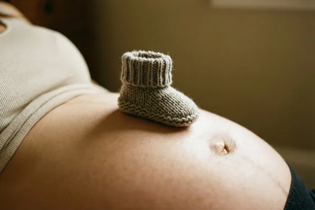 Een klein grijs gebreid babyslofje rust op een ronde zwangere buik in zacht warm licht.