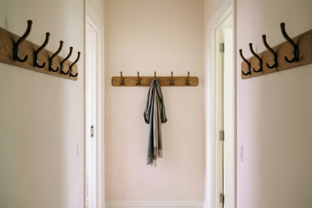 In een gang met lichte muren hangen drie houten kapstokken, waarvan twee leeg zijn en één een grijze sjaal draagt.