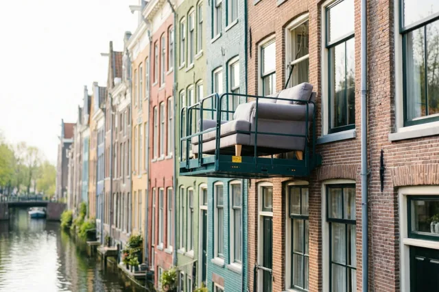 Een meubellift tilt een grijze zetel naar een raam van een bakstenen huis, met kleurrijke huizen langs een kanaal op de achtergrond.