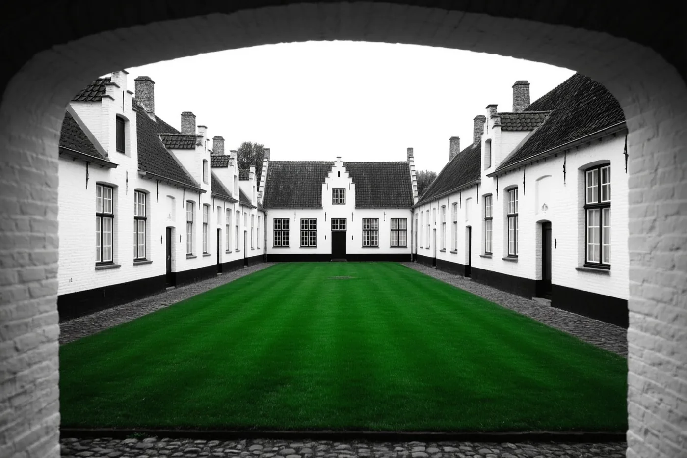 Een bakstenen boog geeft uitzicht op een binnenhof met witgekalkte Vlaamse huizen, waarbij het gras felgroen is en de huizen en lucht in grijstinten zijn weergegeven.