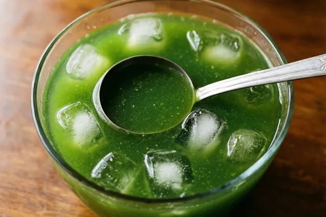 Een glazen kom met groene vloeistof en ijsblokjes staat op een houten ondergrond met een metalen pollepel erin.