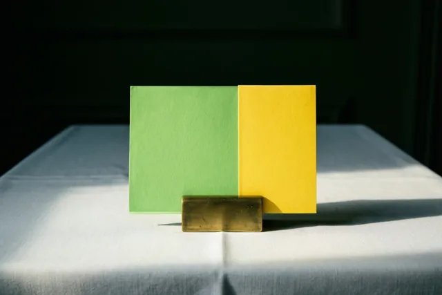 Een groen-geel tafelkaartje staat rechtop in een messing houder op een wit tafelkleed, met scherpe schaduwen door fel zonlicht.