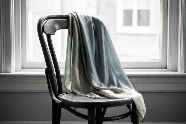 Een zijden sjaal in gedempt groen, blauw en crème hangt over de rug van een donkere gebogen houten stoel bij een helder raam met witte kozijnen.