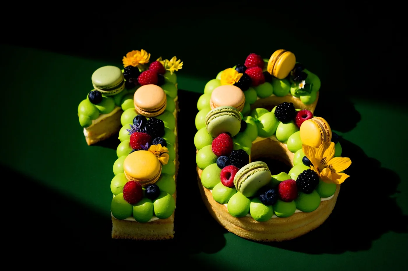 Een taart in de vorm van 16 met groen glazuur, bessen, macarons en eetbare bloemen staat tegen een donkergroen oppervlak in sterk contrasterend licht.