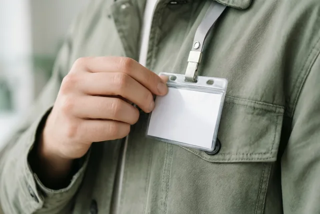 Een hand bevestigt een blanco naambadge aan een groene jas met een halskoord, bij zacht licht en een wazige achtergrond.
