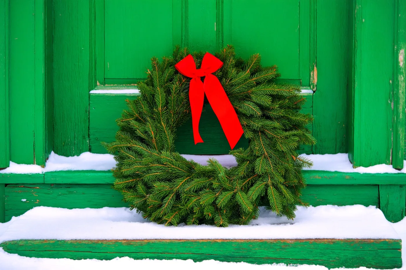 Een groene kerstkrans met een rood lint ligt op een besneeuwde trede voor een oude groene houten deur, met verse sneeuw op de treden en het deurkozijn.