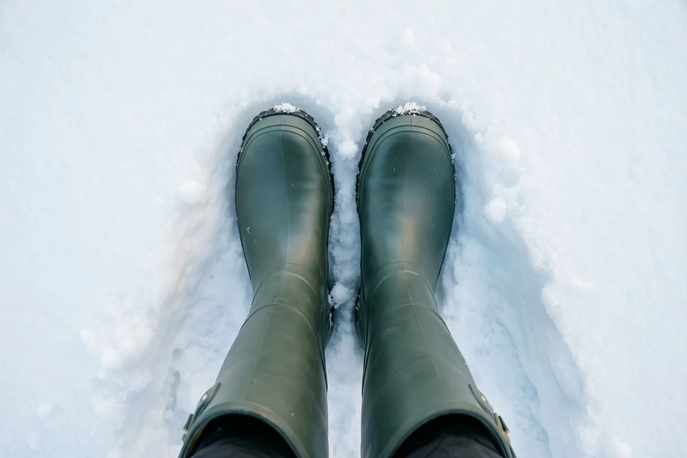 Een paar donkergroene rubberlaarzen staat rechtop in diepe witte sneeuw, van bovenaf gefotografeerd.