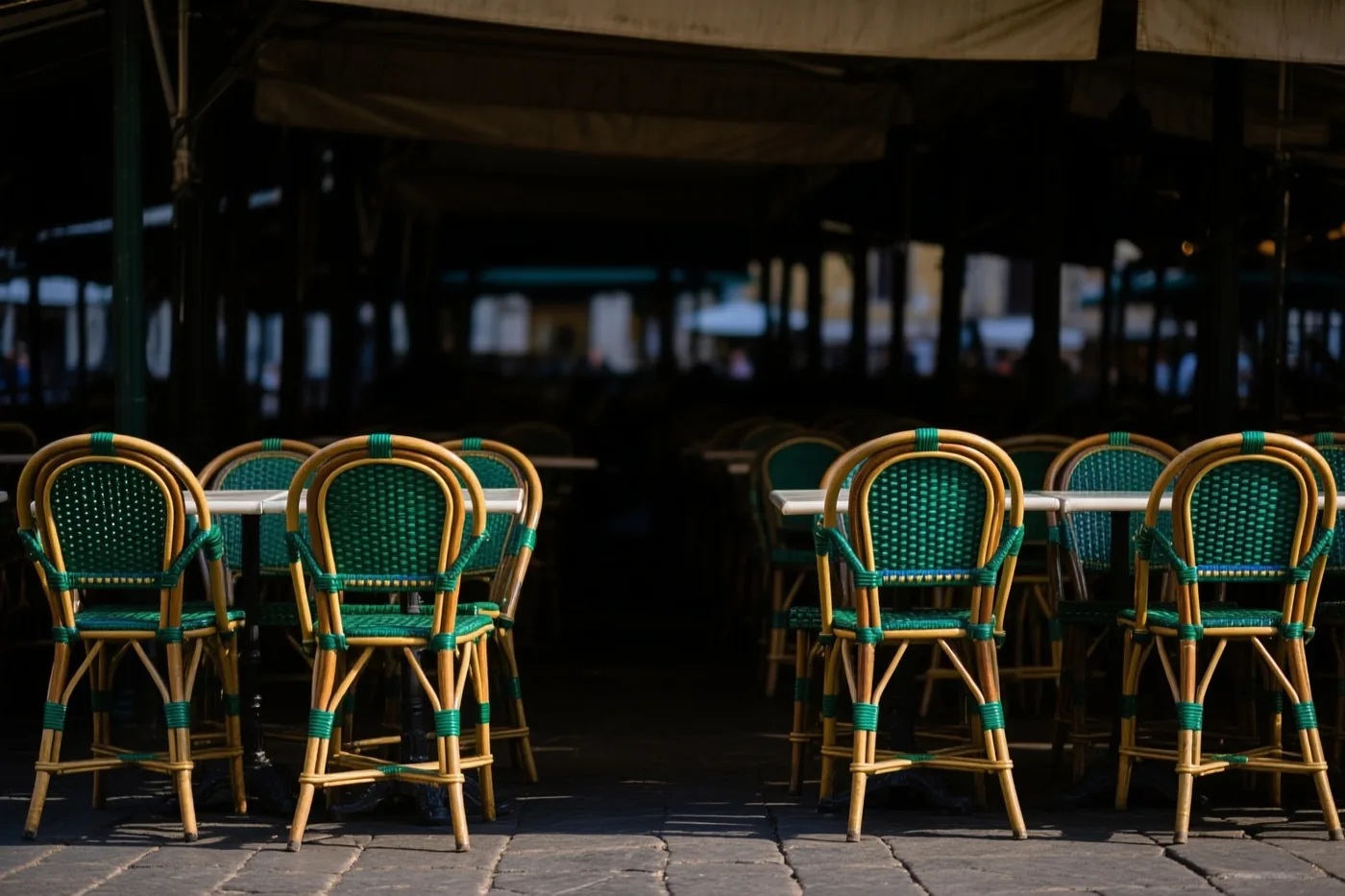 Groene stoelen en bamboestoelen staan rond witte tafels op een stenen caféterras onder een overkapping in warm zonlicht.