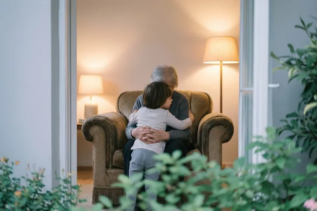Een klein kind zit van achteren gezien tegen een grootouder aan in een grote fauteuil bij warm lamplicht, in een interieur met zachte beige, grijze en groene tinten.