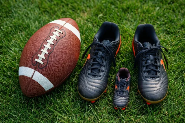 Een bruine Americanfootballbal met witte strepen ligt op groen gras naast een groot en een klein paar zwarte sportschoenen met oranje accenten.