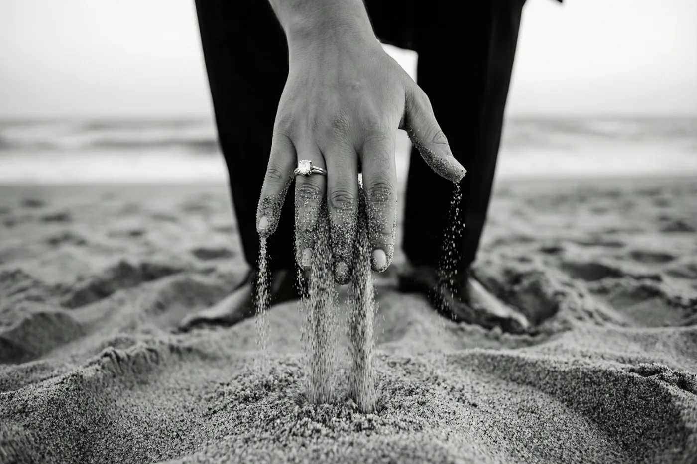Een zwart-witfoto toont van dichtbij een hand met een verlovingsring die op het strand zand door de vingers laat glijden.
