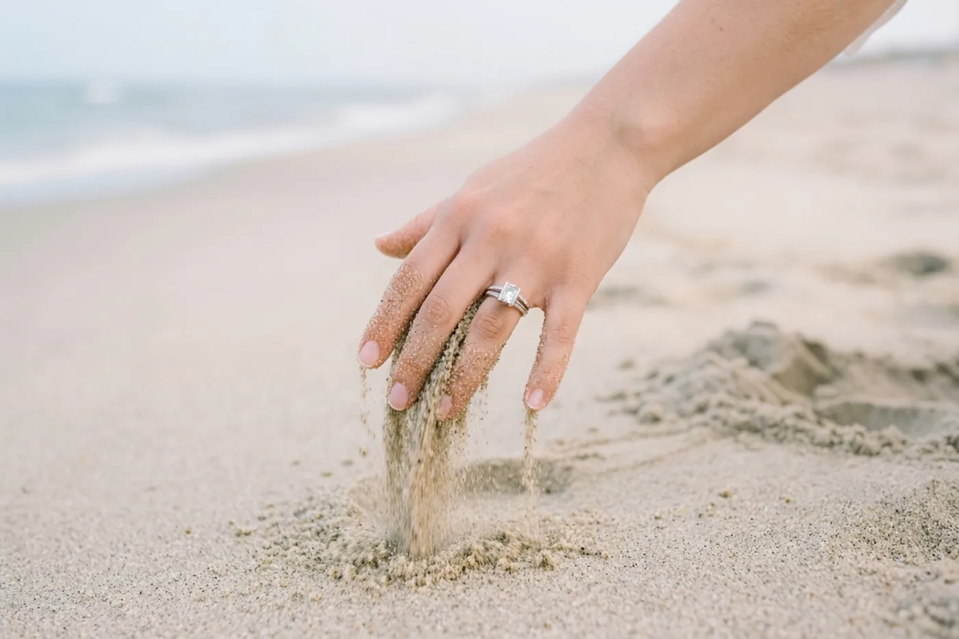 Een hand met een verlovingsring laat zand door de vingers lopen voor een onscherpe zee, bij zacht natuurlijk licht in neutrale tinten.