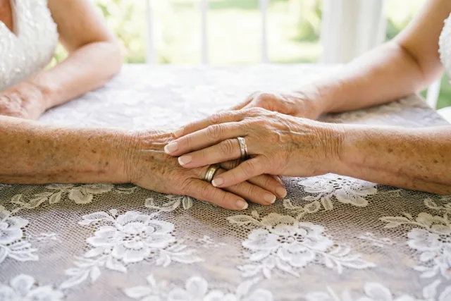Twee oudere handen met trouwringen houden elkaar vast op een wit kanten tafelkleed met bloemenpatroon, in zacht licht van een raam op de achtergrond.