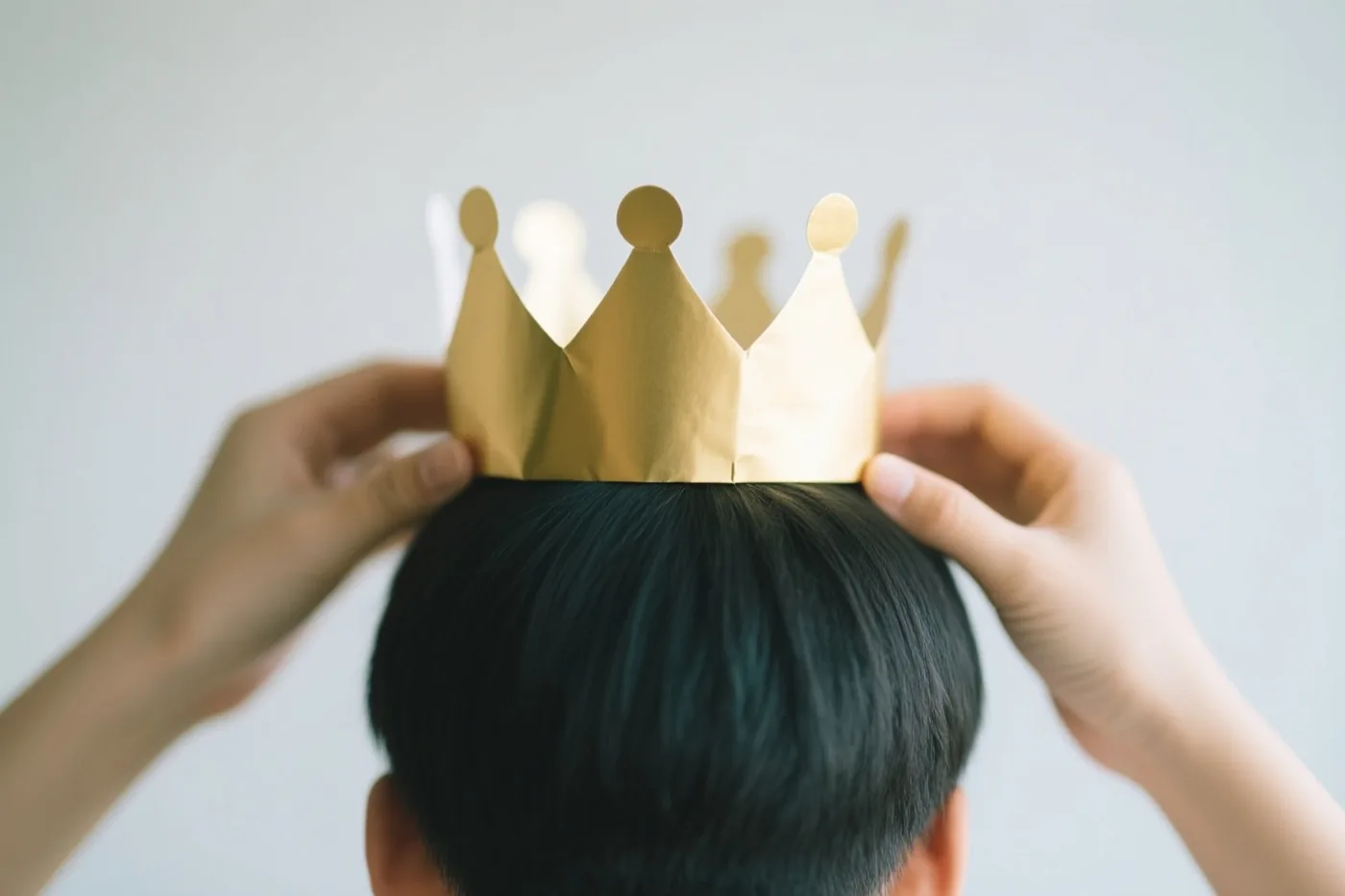 Twee handen plaatsen een goudkleurige papieren kroon op het hoofd van een persoon met donker haar, van achteren gezien tegen een lichtgrijze achtergrond.