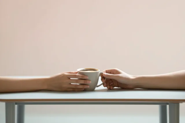 Twee handen houden samen één koffiekop vast boven een tafel tegen een effen achtergrond in zachte pasteltinten.