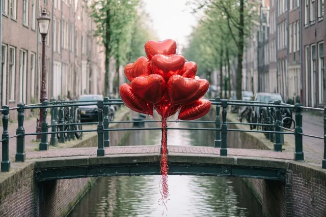 Glanzende rode hartvormige ballonnen hangen aan de leuning van een smalle bakstenen brug over een kanaal, met bakstenen gebouwen en bomen langs het water in zacht daglicht.