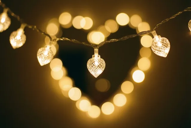 Een slinger hartvormige lichtjes hangt voor een donkere achtergrond, waar wazige lichtcirkels samen een hart vormen in warm goudkleurig licht.