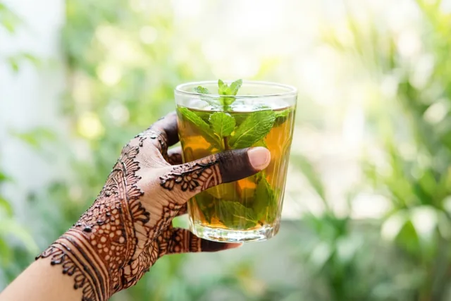 Een hand met fijne hennapatronen houdt een helder glas muntthee met verse groene blaadjes vast tegen een wazige groenwitte achtergrond.