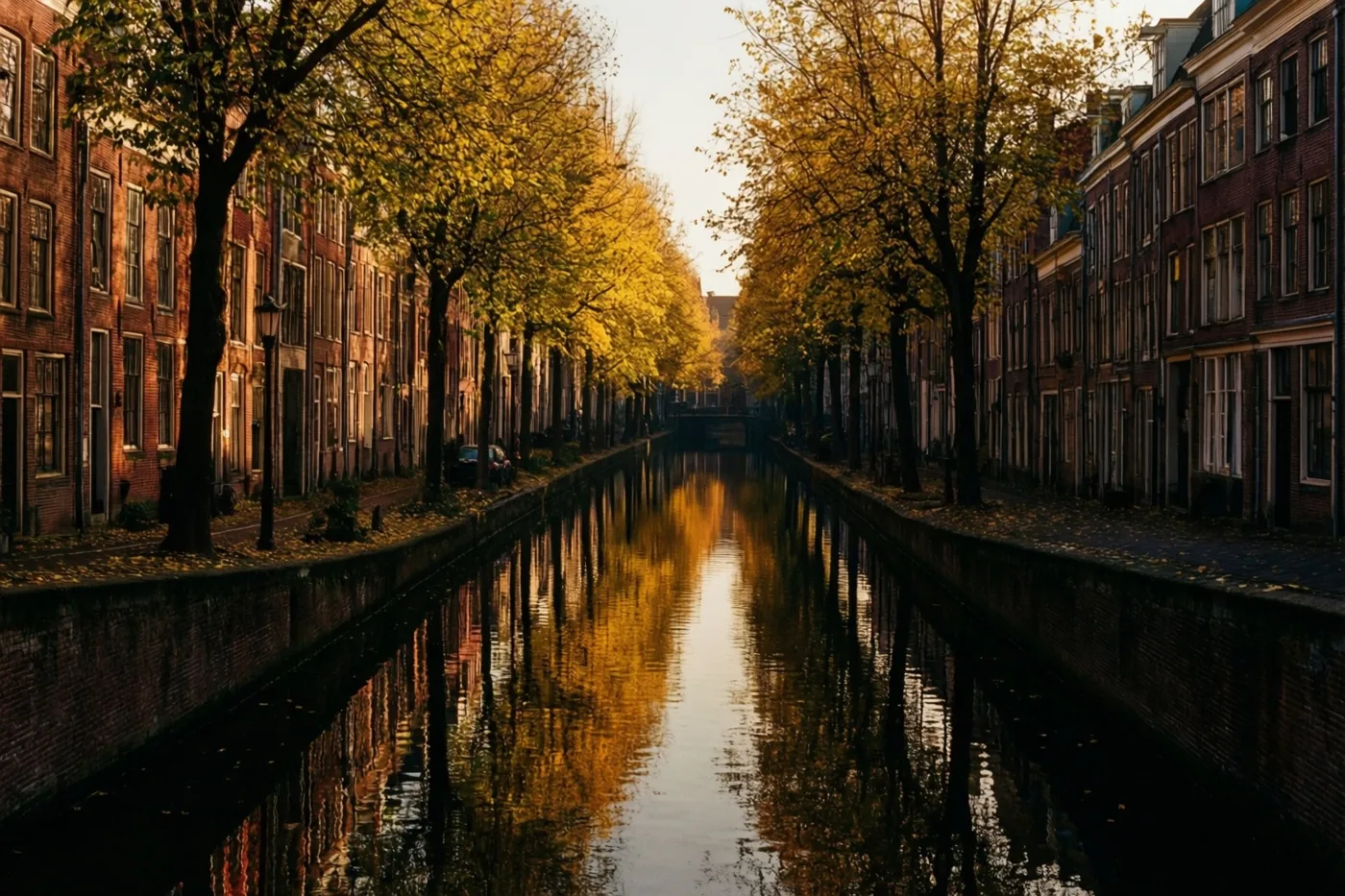 Oude bakstenen gebouwen en goudkleurige herfstbomen staan langs een rustig kanaal en weerspiegelen in het stille water in zacht ochtendlicht.