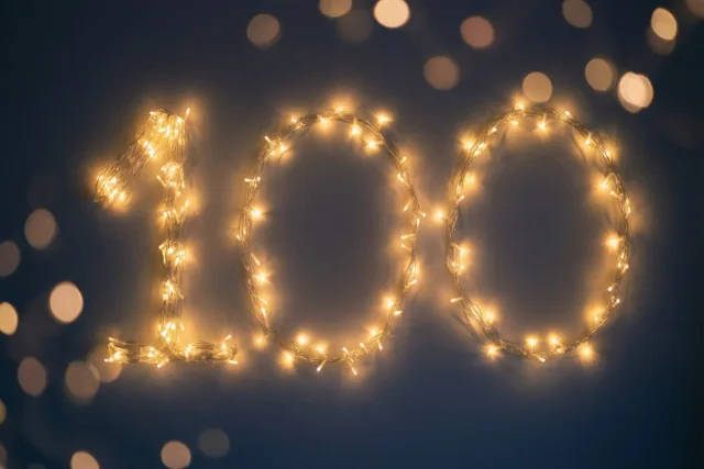 Warme lichtjes vormen het getal 100 tegen een donkere muur, met onscherpe lichtpunten op de achtergrond.