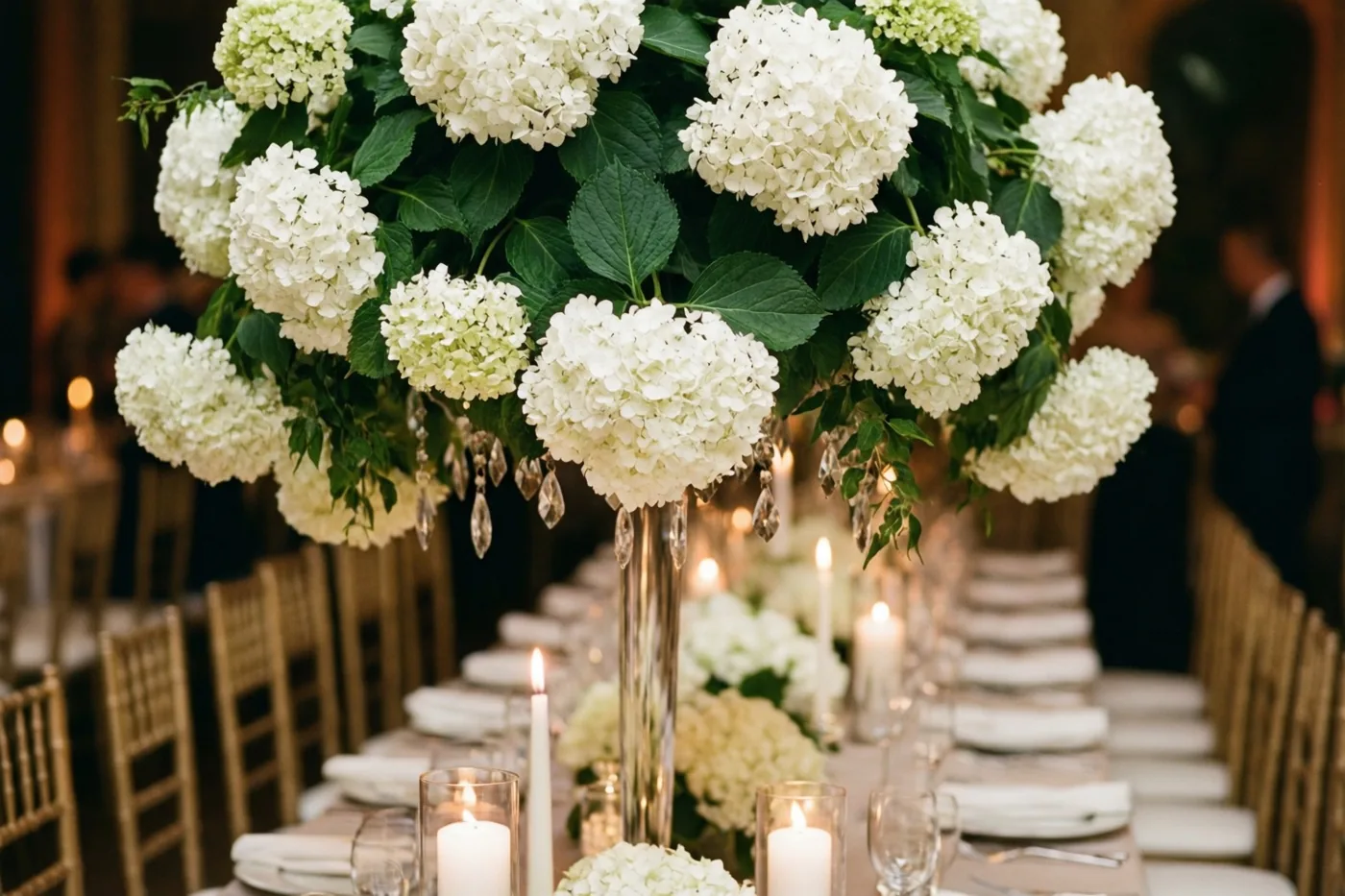 Een lange trouwtafel met witte kaarsen en kristallen glazen heeft in het midden een hoog bloemstuk met afhangende witte hortensia’s, in warm licht.