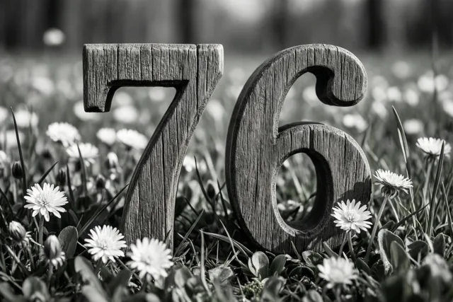 Een zwart-witfoto toont houten cijfers die 76 vormen in een grasweide met kleine witte bloemen.