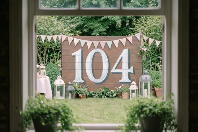 Door een raamkozijn is een groene tuin te zien met een groot houten bord met 104, vlaggetjes en lantaarns in zachte pastel- en aardetinten.