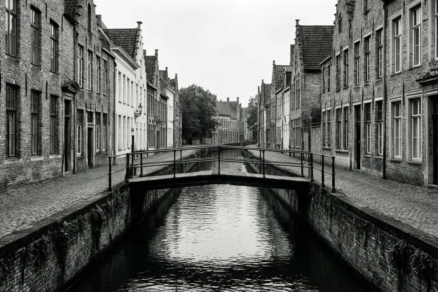 Een monochroom beeld toont een smalle houten brug over een rustig kanaal in een oud Vlaams stadje, met bakstenen gebouwen, puntdaken en hoge ramen erlangs.