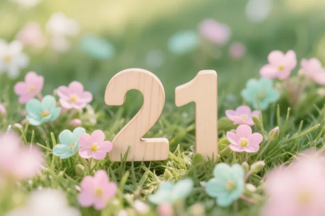 Houten cijfers vormen 21 tussen groen gras en kleine roze en lichtblauwe bloemen in fel zonlicht.
