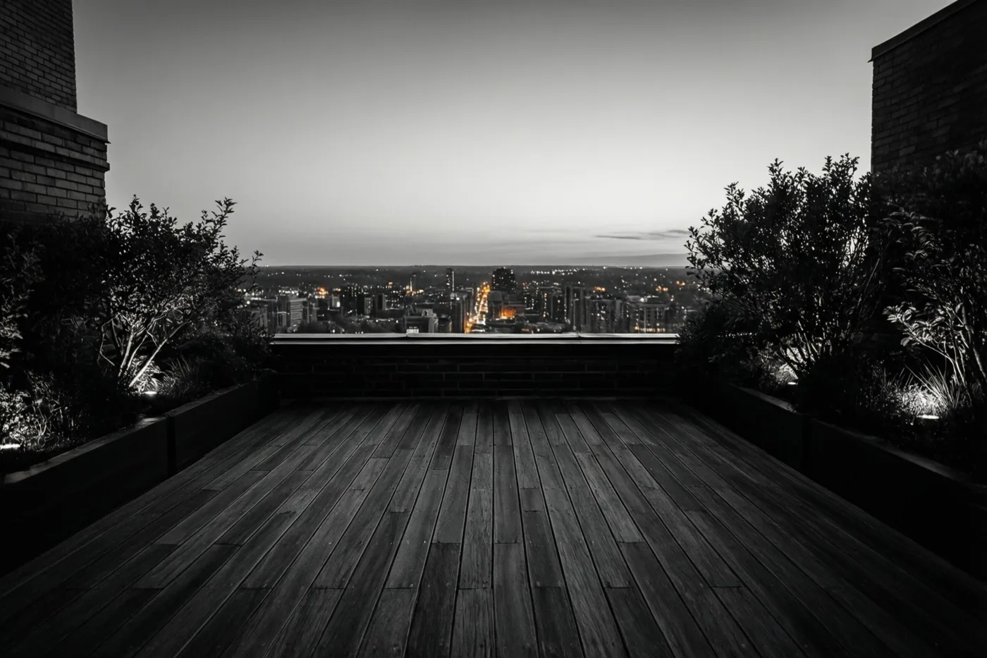 Een dakterras met een houten vloer en planten in potten kijkt bij schemering uit over een verlichte stad, in een monochroom beeld met subtiele warme lichtaccenten.