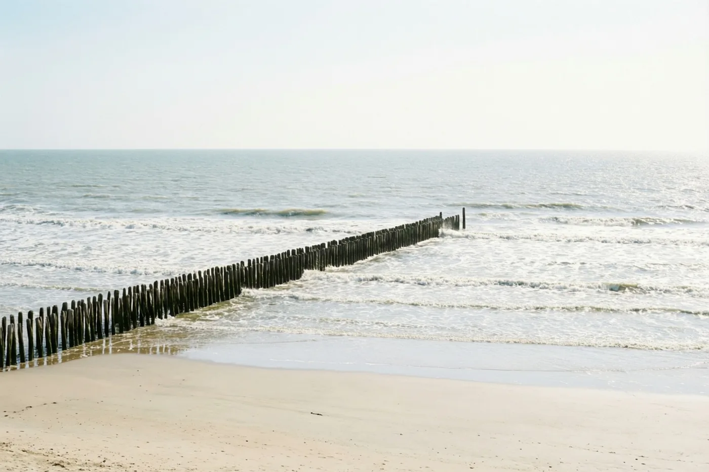 Een lange houten golfbreker loopt van een zandstrand de zee in, met zachte golven onder een bleke hemel in beige, grijze en blauwe tinten.