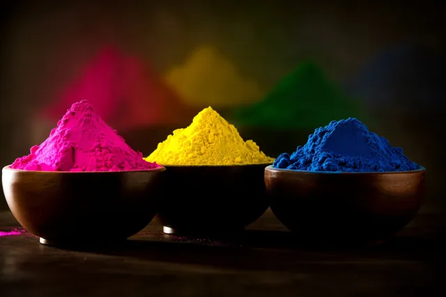 Drie houten kommen met roze, geel en blauw poeder staan op een rij voor een donkere achtergrond, met groen poeder onscherp daarachter.
