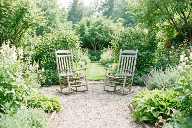 Twee houten schommelstoelen staan op een grindpad tussen bloeiende planten en bomen in een groene tuin, met licht dat door de bladeren valt.