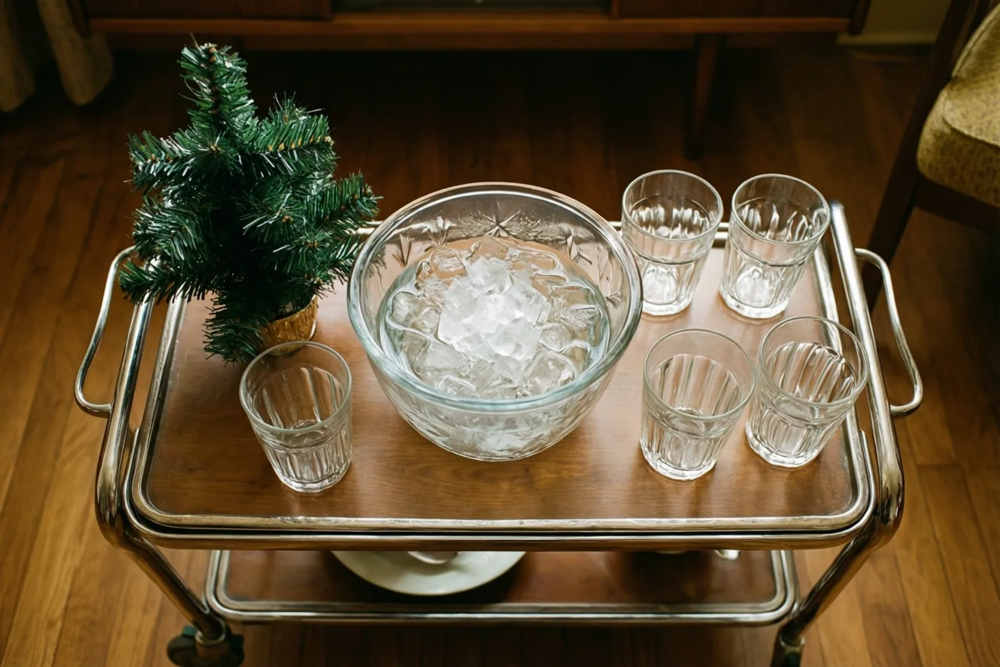 Een houten serveerwagen met chromen handgrepen draagt een glazen punchschaal met ijs, vijf doorzichtige drinkglazen en een klein kerstboompje in een pot, op een houten vloer.
