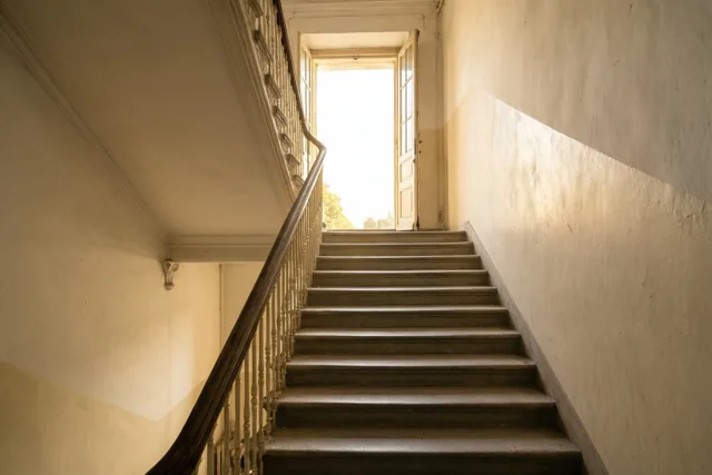 Een houten trap in een oud gebouw loopt omhoog naar een helder verlichte open deur in warm, goudkleurig licht.