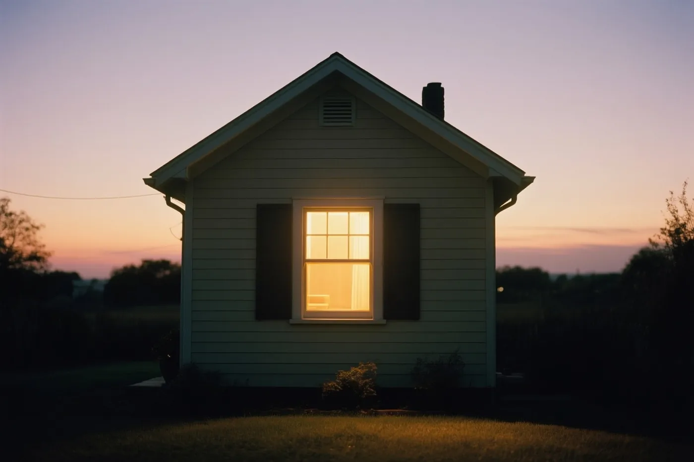 Een klein huis met één verlicht raam staat bij schemering alleen in een grasveld onder een hemel met paarse en oranje tinten.