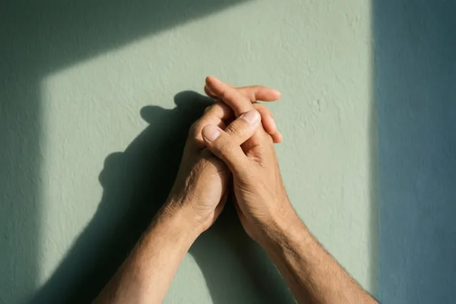 Twee handen houden elkaar vast voor een muur met zichtbare structuur en werpen in het zonlicht een duidelijke schaduw op de muur.