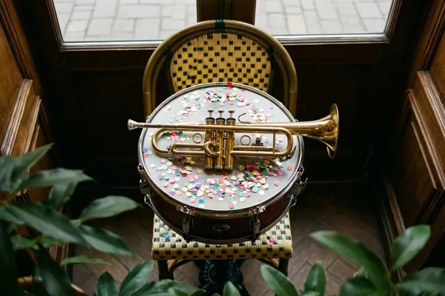 Een gouden trompet ligt op een kleine trom met kleurrijke confetti op een gevlochten caféstoel, binnen bij een houten deur met een raam en groen eromheen.