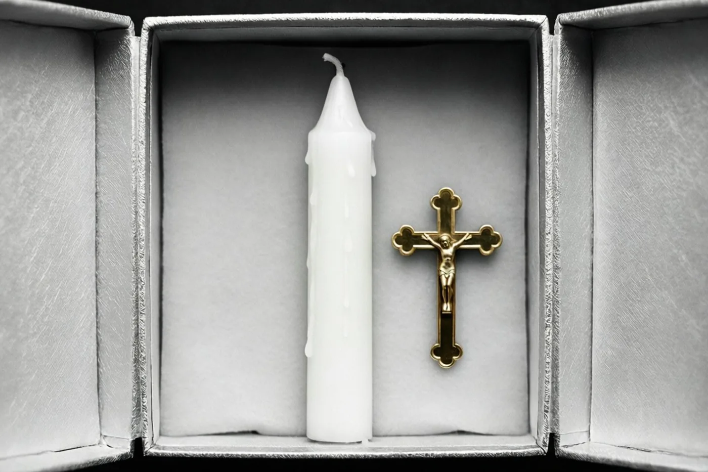 Een open zilveren geschenkdoos bevat een witte communiekaars en een klein gouden kruisje op een witte zachte voering, tegen een zwarte achtergrond.